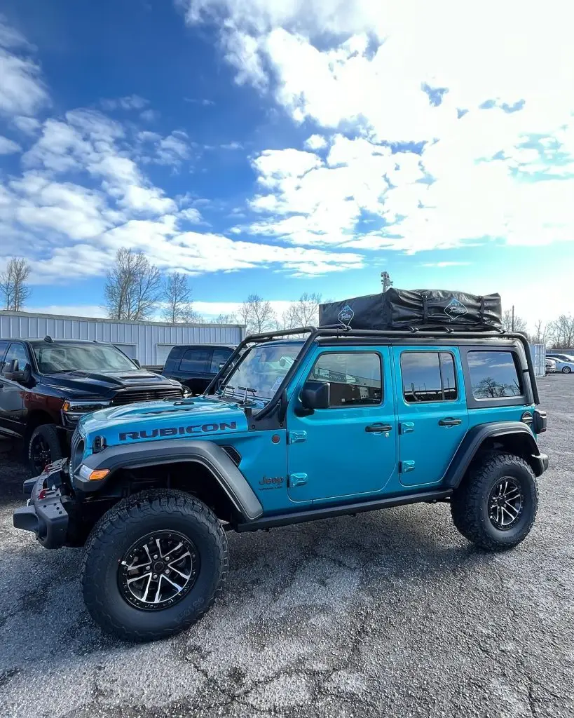 Blue Jeep Rubicon parked under a cloudy sky, equipped with a rooftop tent, showcasing off-road capability.