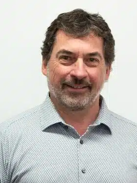 Man with gray hair and beard wearing a patterned shirt, smiling slightly.