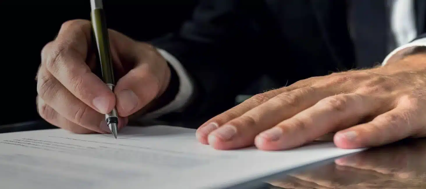 Person signing a document with a pen on a reflective surface, indicating business or legal activity.
