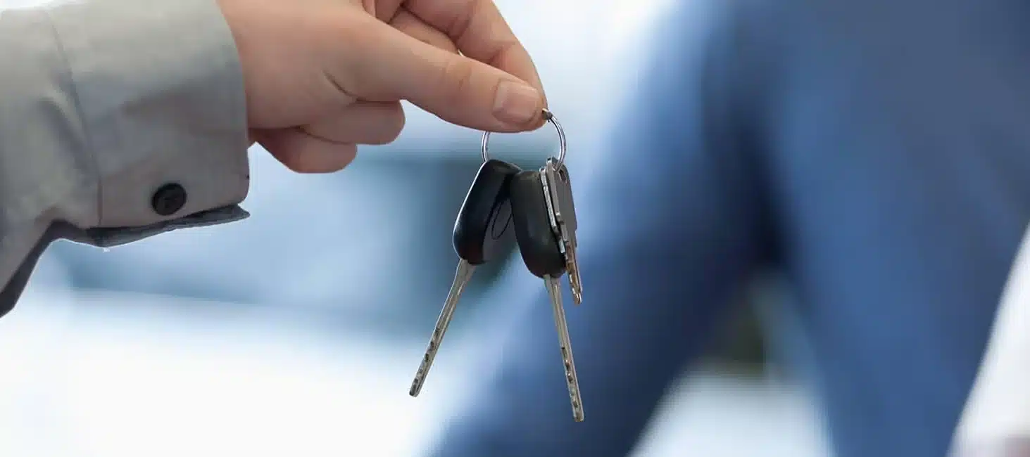 Hand holding car keys against a blurred background, emphasizing vehicle ownership or sale.