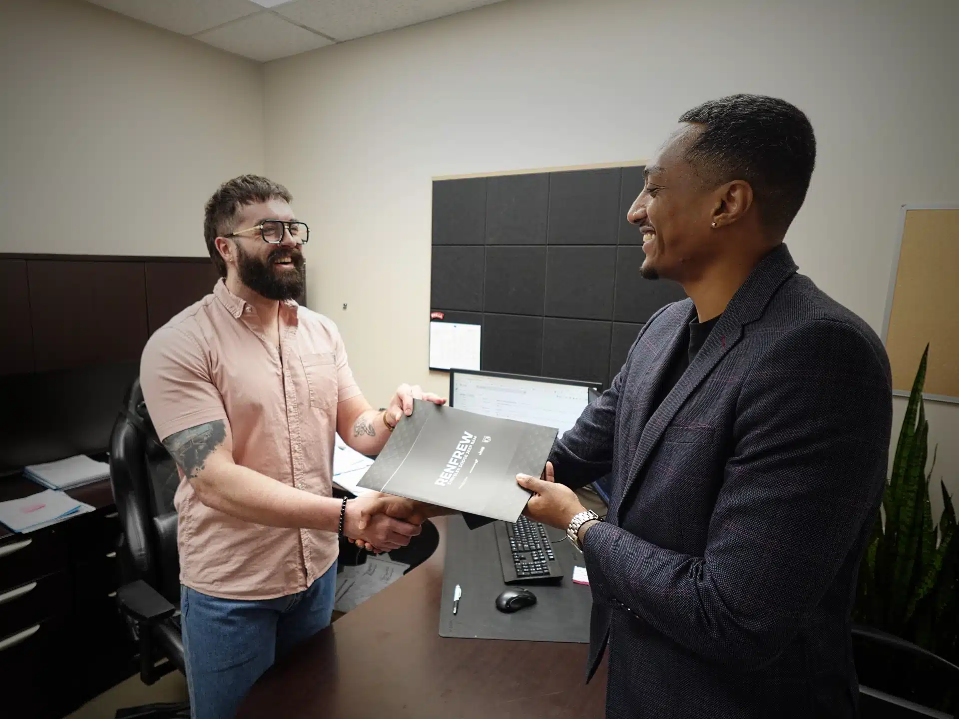 Two colleagues exchange a "Renew" folder in an office, smiling at each other professionally.